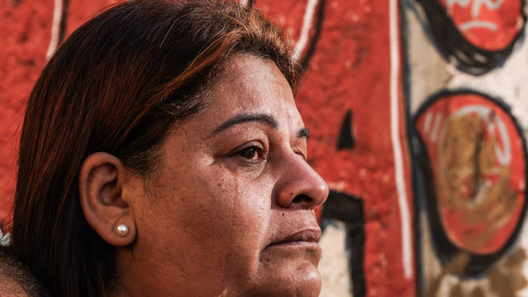 L'immagine mostra il profilo di una donna con i capelli castani lisci, che indossa orecchini di perle. Ha un'espressione seria e sembra che si trovi in un momento di riflessione o tristezza. Lo sfondo è un muro decorato con graffiti colorati, prevalentemente nei toni del rosso e del nero, che aggiungono contrasto alla scena.
