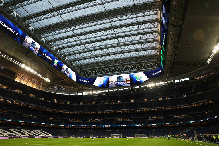 A imagem mostra o interior do Estádio Santiago Bernabéu, casa do Real Madrid. O teto é retrátil e há telões visíveis nas laterais. O campo de futebol está preparado para um jogo, com jogadores em campo e as arquibancadas vazias. A iluminação do estádio é visível, destacando a estrutura moderna do local.