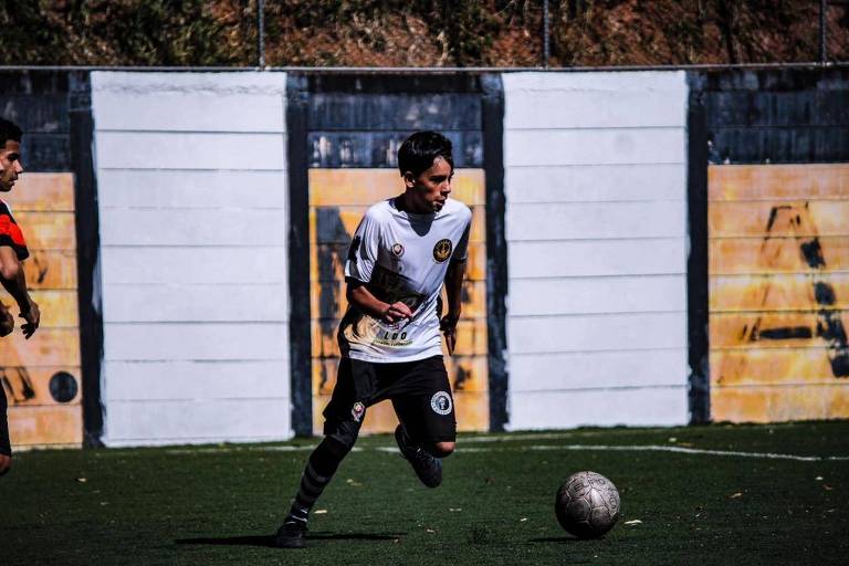 Un giovane calciatore corre attraverso un campo erboso, con un pallone da calcio davanti a sé. Indossa una camicia bianca con dettagli neri e un distintivo sul petto. Sullo sfondo c'è un muro con graffiti gialli e neri. Un altro giocatore, sulla sinistra, è parzialmente visibile: indossa una maglia rossa.
