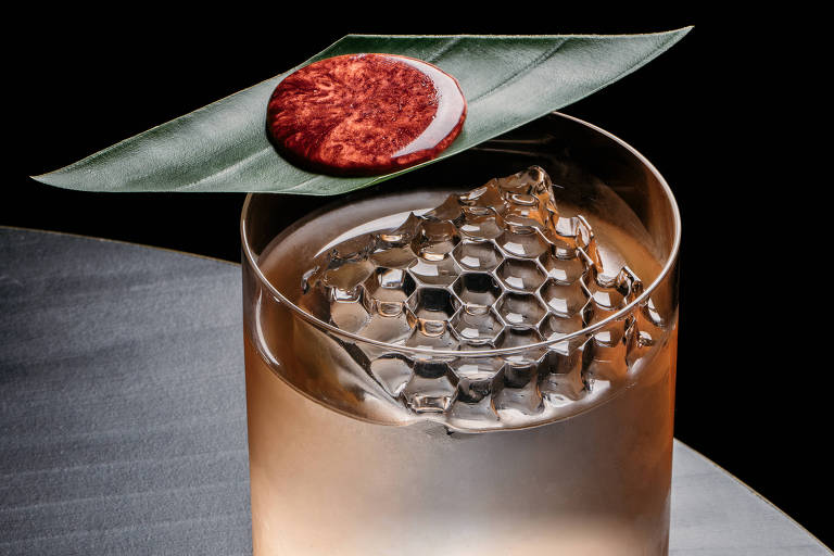A imagem mostra um copo de bebida com gelo em formato de favo de mel, colocado sobre uma mesa escura. Em cima do copo, há uma folha verde com um círculo vermelho brilhante, que parece ser uma decoração ou um ingrediente. O fundo da imagem é preto, destacando o copo e os elementos decorativos.