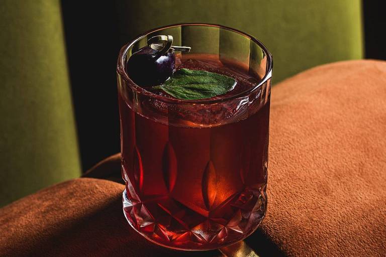 A imagem mostra um coquetel em um copo de cristal, com uma cor vermelha escura. O coquetel é decorado com uma folha de hortelã e uma fruta, possivelmente uma cereja, no topo. O copo está posicionado sobre um sofá de cor laranja, enquanto ao fundo há um tecido verde que cria um contraste. A iluminação destaca a transparência do líquido e os detalhes do copo.
