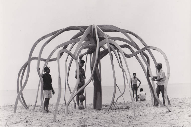 A imagem mostra uma estrutura artística em forma de árvore ou planta, com longos ramos curvados, feita de material claro. Há cinco pessoas ao redor da estrutura, algumas segurando ferramentas, enquanto outras observam. O cenário é uma praia, com areia e o mar ao fundo, em um dia claro.
