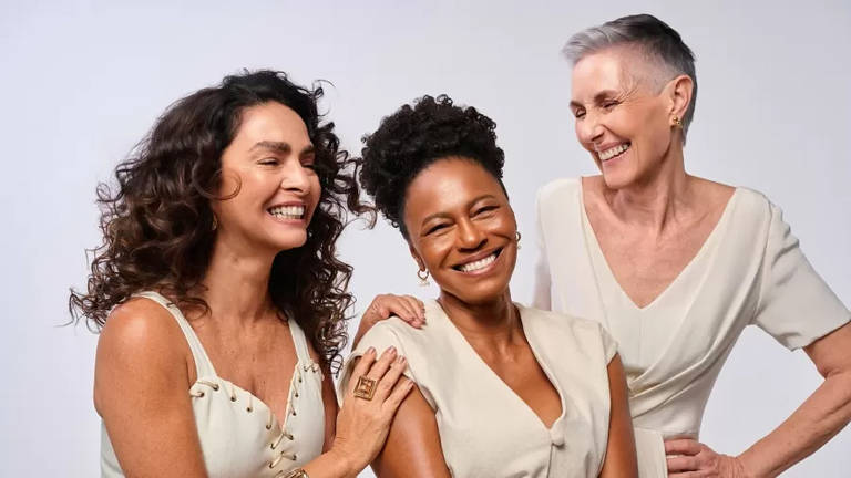 A imagem mostra três mulheres sorrindo e se divertindo juntas. Elas estão vestidas com roupas claras e têm diferentes estilos de cabelo. A mulher à esquerda tem cabelo cacheado e usa uma blusa com detalhes. A mulher no centro tem cabelo preso e um sorriso largo, vestindo uma blusa clara. A mulher à direita tem cabelo curto e grisalho, usando uma blusa com decote em V. Todas parecem felizes e estão em um ambiente neutro.