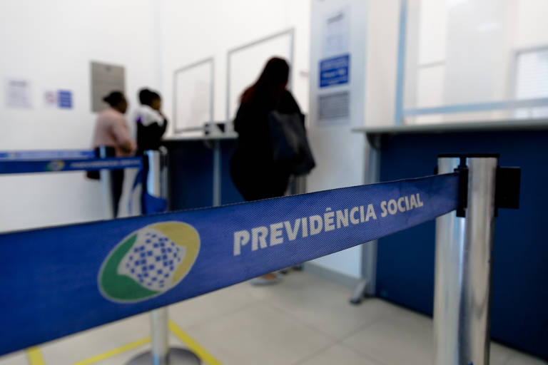 A imagem mostra o interior de uma agência da Previdência Social, onde algumas pessoas estão em fila. No primeiro plano, há uma fita de isolamento com a inscrição 'PREVIDÊNCIA SOCIAL'. Ao fundo, é possível ver paredes brancas e janelas, além de pessoas aguardando atendimento.