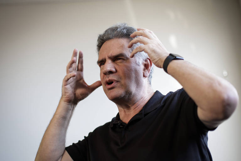 Um homem de cabelos grisalhos e curtos, vestindo uma camiseta preta, está em uma pose expressiva, com as mãos levantadas ao lado da cabeça, como se estivesse enfatizando um ponto durante uma conversa. O fundo é neutro e claro, sugerindo um ambiente interno.