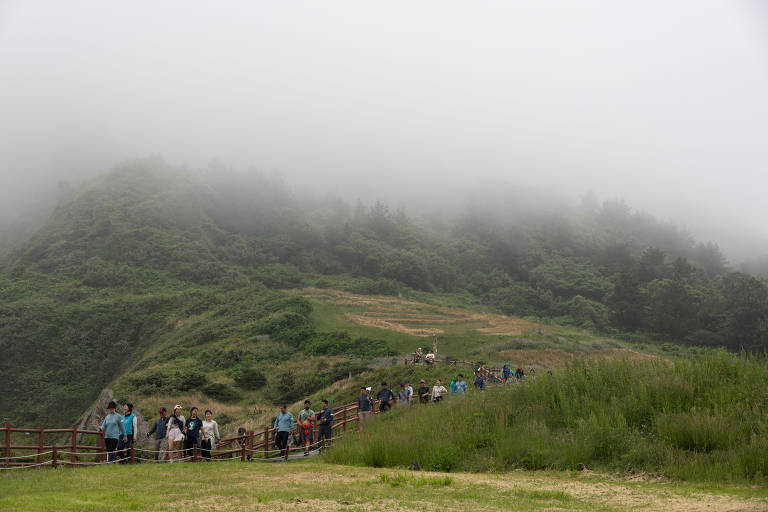 A imagem mostra um grupo de pessoas caminhando em uma trilha em uma montanha coberta por névoa. O terreno é verde e há uma cerca de madeira ao longo do caminho. Ao fundo, a montanha é visível, mas parcialmente encoberta pela névoa. O clima parece nublado e úmido, com vegetação densa ao redor.