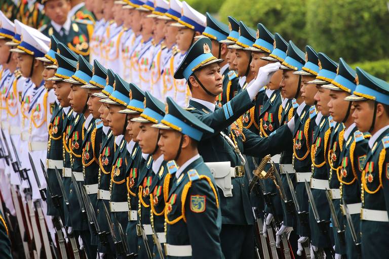 A imagem mostra uma formação de soldados em trajes de gala, alinhados em uma cerimônia militar. Os soldados estão em posição de sentido, com uniformes escuros e chapéus com detalhes em azul. Um dos soldados, em destaque, está levantando a mão em um gesto de saudação. Ao fundo, há outros soldados em uniformes brancos, formando uma segunda fileira. O ambiente parece ser ao ar livre, com vegetação ao fundo.