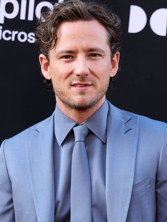 Um homem com cabelo cacheado e curto, usando um terno azul claro e uma gravata. Ele está posando para a foto em um evento, com um fundo preto que contém logotipos. O homem tem uma expressão neutra e está olhando diretamente para a câmera.