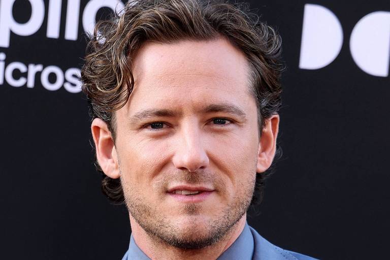 Um homem com cabelo cacheado e curto, usando um terno azul claro e uma gravata. Ele está posando para a foto em um evento, com um fundo preto que contém logotipos. O homem tem uma expressão neutra e está olhando diretamente para a câmera.
