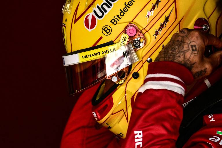 A imagem mostra um piloto de Fórmula 1 usando um capacete amarelo com detalhes vermelhos e logotipos. O piloto está ajustando o capacete, que possui uma viseira transparente. O fundo é de cor escura, destacando o capacete e a roupa vermelha do piloto. É possível ver uma tatuagem na mão do piloto.