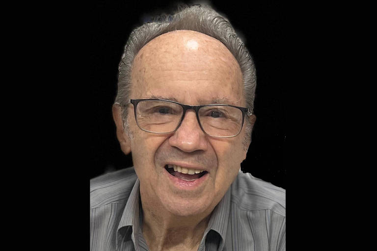 Um homem idoso com óculos e cabelo grisalho, sorrindo. Ele está usando uma camisa de botão cinza e o fundo da imagem é preto, destacando seu rosto e expressão amigável.