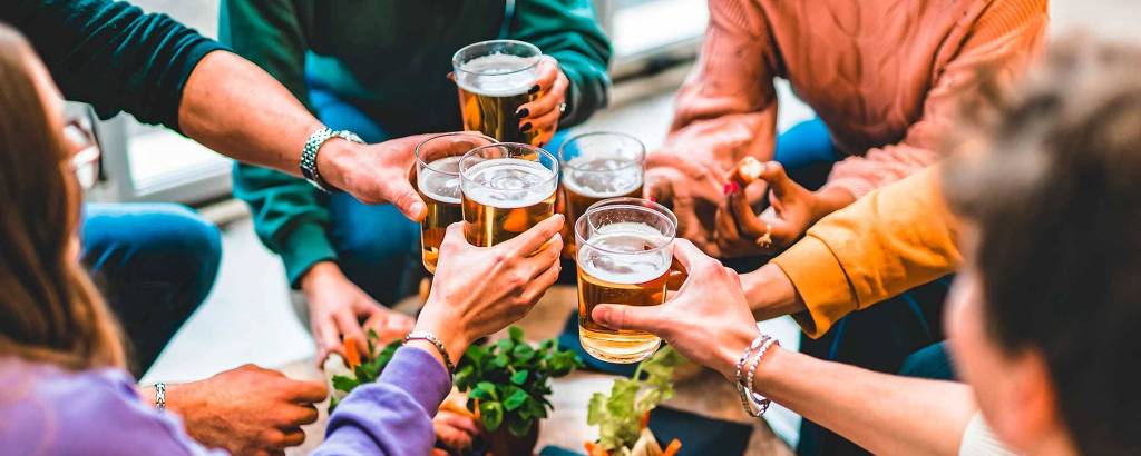Um grupo de pessoas está reunido em torno de uma mesa, levantando copos de cerveja em um brinde. A mesa está cheia de petiscos variados, como carnes e vegetais. As pessoas estão sorrindo e se divertindo, vestindo roupas casuais. O ambiente parece ser descontraído e acolhedor, com luz natural entrando pelas janelas ao fundo.