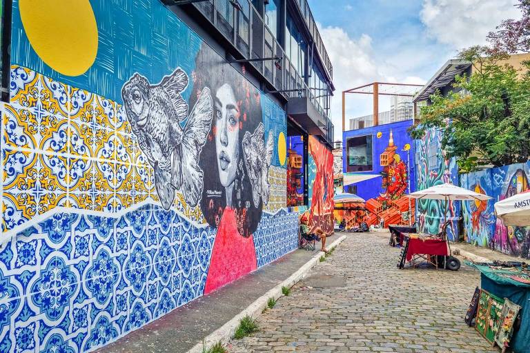 A imagem mostra uma rua estreita cercada por murais coloridos em paredes de edifícios. Os murais apresentam uma variedade de estilos, incluindo padrões azulejados, retratos e elementos gráficos vibrantes. Há mesas e cadeiras ao ar livre, além de uma sombrinha branca. O céu está parcialmente nublado, e a rua é pavimentada com pedras.