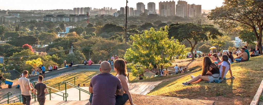 A imagem mostra um parque urbano ao entardecer, com várias pessoas sentadas e relaxando em um gramado. Ao fundo, há uma vista da cidade com prédios altos. Árvores e vegetação estão presentes, e a luz do sol cria um ambiente acolhedor. Algumas pessoas estão em grupos, enquanto outras estão sozinhas, aproveitando o momento.