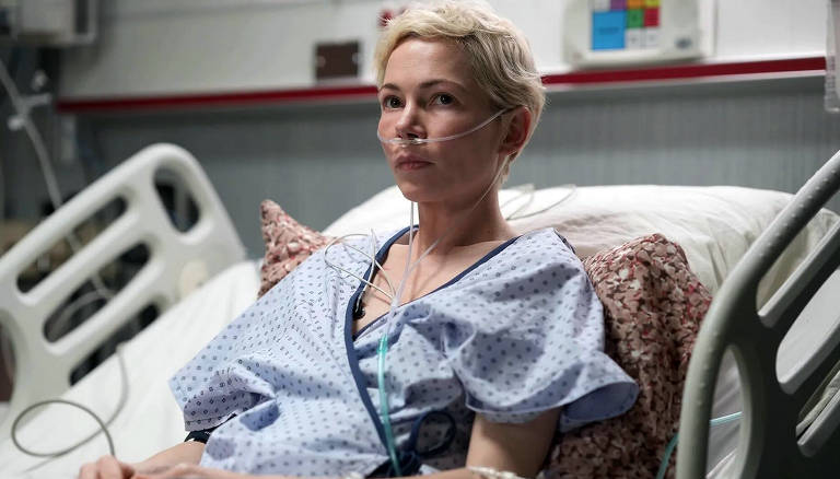 A imagem mostra uma mulher sentada em uma cama de hospital. Ela tem cabelo curto e platinado, e está usando um pijama hospitalar azul com pequenos padrões. A mulher está conectada a tubos de oxigênio que passam pelo seu nariz. O ambiente ao redor é típico de um hospital, com equipamentos médicos visíveis ao fundo e uma cama com lençóis brancos e uma almofada decorativa.la tem cabelo curto e platinado, e está usando um pijama hospitalar azul com pequenos padrões. A mulher está conectada a tubos de oxigênio que passam pelo seu nariz. O ambiente ao redor é típico de um hospital, com equipamentos médicos visíveis ao fundo e uma cama com lençóis brancos e uma almofada decorativa.