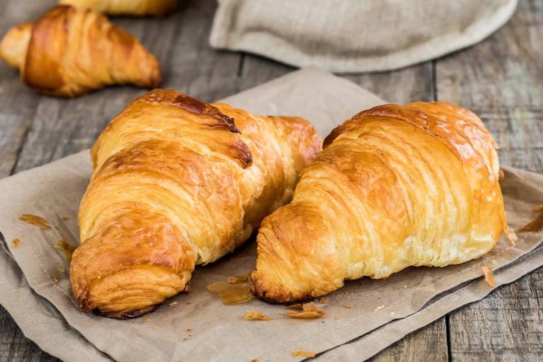 A imagem mostra croissants frescos dispostos sobre uma superfície de madeira. Dois croissants grandes estão em destaque sobre um papel pardo, enquanto outros croissants menores estão em um saco de tecido ao fundo. A iluminação é suave, destacando a textura dourada e folhada dos croissants.