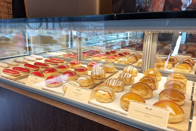 A imagem mostra uma vitrine de doces em uma confeitaria. Há uma variedade de doces dispostos em fileiras, incluindo bolos com coberturas brilhantes, como geléias vermelhas e douradas, além de doces com drapeados de chocolate. Os doces estão organizados em uma bandeja branca, e algumas etiquetas com nomes estão visíveis na parte inferior.