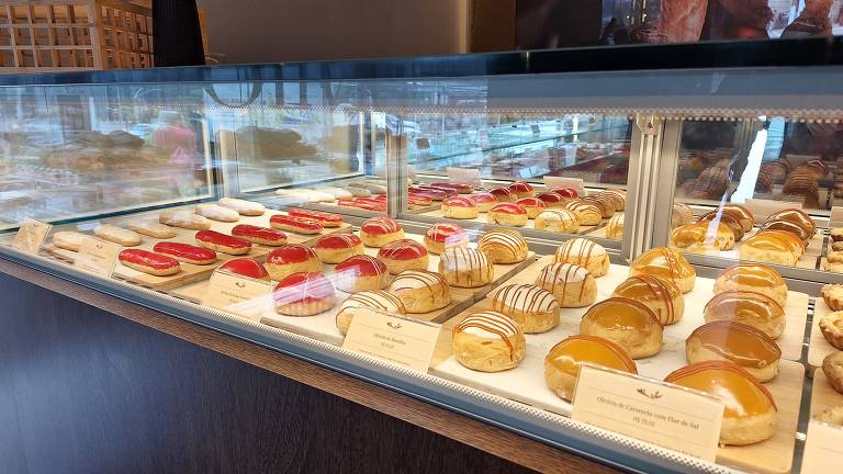 A imagem mostra uma vitrine de doces em uma confeitaria. Há uma variedade de doces dispostos em fileiras, incluindo bolos com coberturas brilhantes, como geléias vermelhas e douradas, além de doces com drapeados de chocolate. Os doces estão organizados em uma bandeja branca, e algumas etiquetas com nomes estão visíveis na parte inferior.
