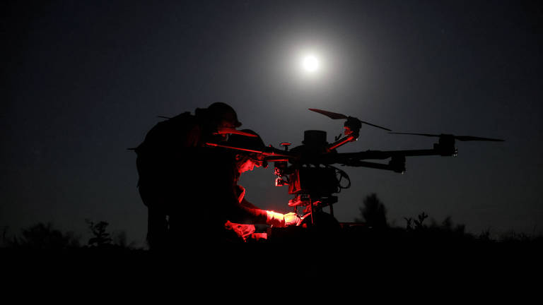 A imagem mostra duas silhuetas de pessoas trabalhando em um drone sob a luz da lua cheia. Uma das pessoas está agachada, iluminando algo com uma luz vermelha, enquanto a outra está em pé, observando. O céu está escuro, e a lua brilha intensamente ao fundo, criando um contraste com as figuras em primeiro plano.