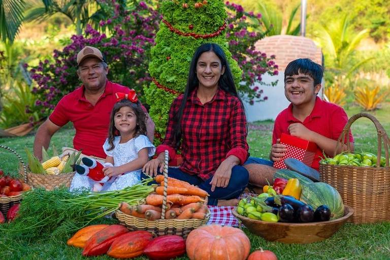 Uma família composta por um homem, uma mulher, uma menina e um menino está sentada em um gramado verde. Eles estão cercados por cestas cheias de frutas e vegetais, incluindo tomates, cenouras, abóbora, bananas e outros produtos. Ao fundo, há uma árvore ornamental e plantas floridas, além de uma estrutura branca que parece ser uma churrasqueira. Todos estão sorrindo e vestindo roupas casuais, com a mulher usando uma blusa xadrez e os homens camisetas vermelhas.