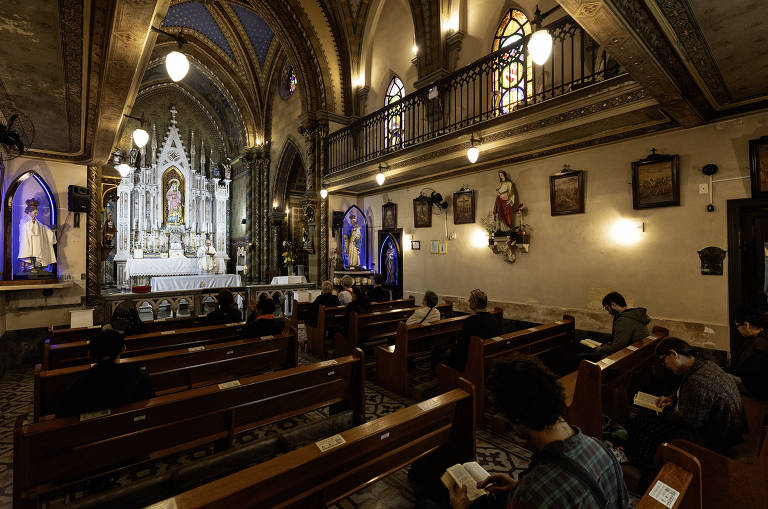 A imagem mostra o interior de uma igreja com um altar ao fundo, decorado com estátuas e elementos religiosos. Há bancos de madeira ocupados por pessoas em oração. O teto é alto e apresenta detalhes arquitetônicos, com iluminação suave proveniente de lâmpadas pendentes. As paredes são adornadas com quadros e estátuas. A atmosfera é de tranquilidade e devoção