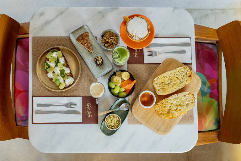 Uma mesa de café da manhã com uma variedade de alimentos dispostos. Há um prato redondo com torradas cobertas com abacate e fatias de limão, um prato retangular com pães, uma tigela com granola, frutas como banana e limão, e um copo com uma bebida. A mesa é de mármore e as cadeiras têm almofadas coloridas.