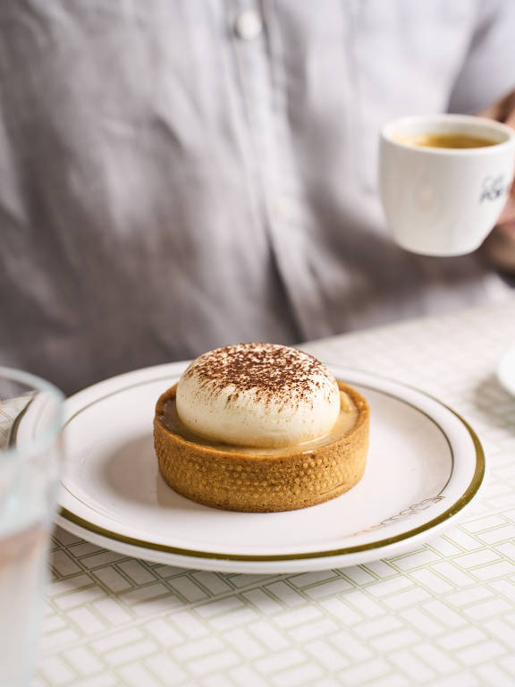A imagem mostra um prato com uma sobremesa em destaque, que é uma torta com um topo cremoso polvilhado com cacau. Ao lado, há uma xícara de café e um copo de água. O fundo é uma mesa com uma toalha de padrão geométrico. A mão de uma pessoa segurando a xícara de café é visível.
