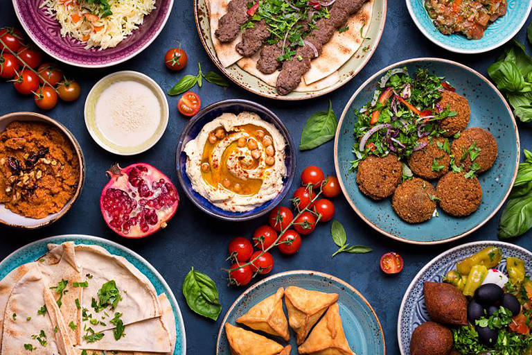 A imagem mostra uma mesa repleta de pratos variados de comida. Há uma porção de arroz, kebabs grelhados, pães, homus com grão-de-bico, falafel, salada, e uma variedade de molhos e acompanhamentos. Também estão presentes tomates-cereja, uma romã cortada, e pastéis em formato triangular. Os pratos são coloridos e decorados com ervas frescas, criando uma apresentação vibrante e apetitosa.