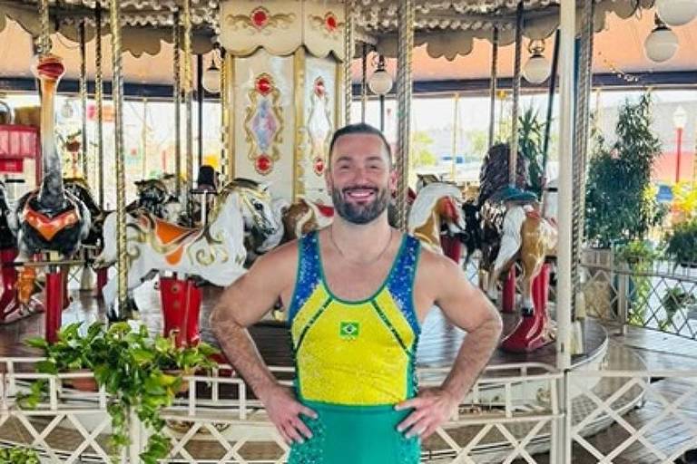 Um homem está posando em um carrossel. Ele usa um traje de cores verde e amarelo, que remete à bandeira do Brasil, e está com um sorriso no rosto. O fundo mostra vários cavalos de carrossel, alguns decorados, e uma estrutura de madeira. O ambiente é iluminado e parece ser um local de diversão.