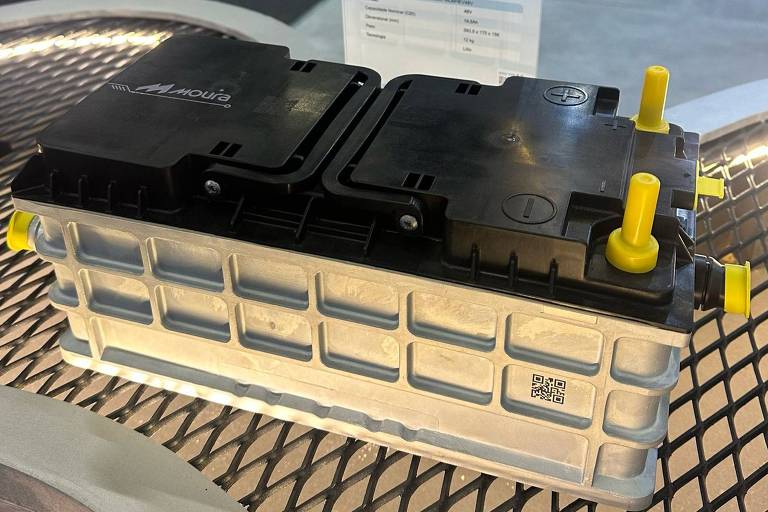 A imagem mostra uma bateria automotiva com um design moderno. A bateria é composta por uma parte inferior em plástico claro e uma parte superior preta, com dois conectores amarelos visíveis. Ao fundo, há uma placa informativa que parece descrever as especificações do produto, mas o texto não é legível na imagem.