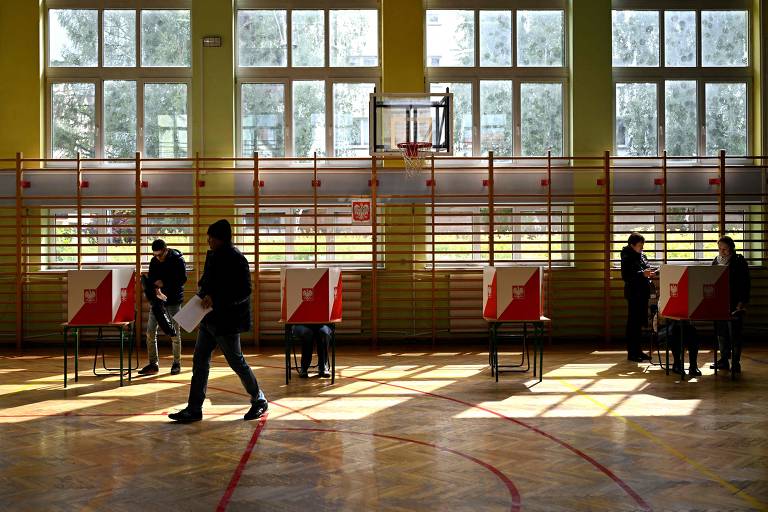 A imagem mostra um ambiente interno de uma escola, onde várias pessoas estão participando de uma votação. Há cabines de votação vermelhas e brancas dispostas em fileiras, e algumas pessoas estão em pé, enquanto outras estão sentadas. A luz do sol entra pelas janelas grandes, iluminando o espaço. Ao fundo, há uma quadra de esportes com uma tabela de basquete.