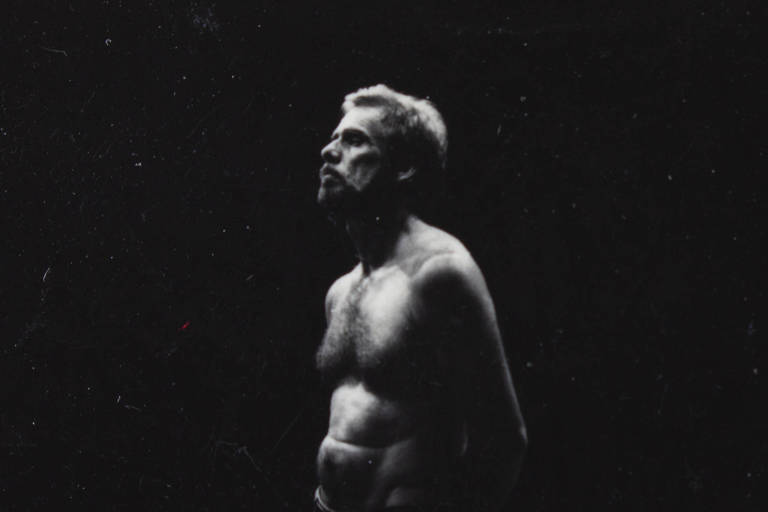 L'image montre un homme debout, torse nu et cheveux courts, le regard tourné vers le haut. L'éclairage est saisissant, mettant en valeur son visage et une partie de son corps, tandis que l'arrière-plan est sombre. L'homme a une expression sérieuse et contemplative, et l'image présente un contraste élevé, avec une prédominance de tons sombres.