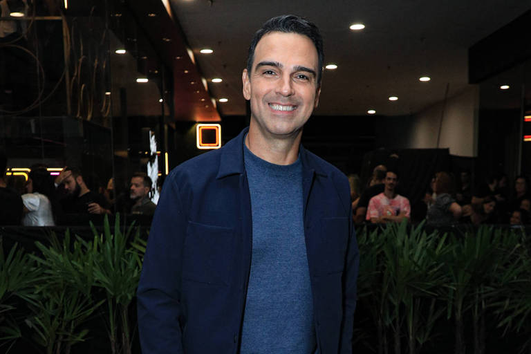 Um homem está posando para a foto em um evento social. Ele está vestido com uma blusa azul escura e um blazer combinando, além de calças escuras. O homem sorri e está de pé em um ambiente interno, com plantas ao fundo e uma multidão visível. O piso é de concreto e a iluminação é suave.