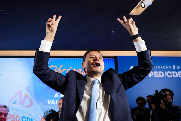 Um homem em um terno escuro e gravata clara levanta os braços em sinal de celebração, fazendo o gesto de vitória com as mãos. Ele está sorrindo e parece estar em um evento político, com um fundo que inclui logotipos de partidos e uma tela com a palavra 'Obrigado'. Há pessoas ao fundo, algumas com câmeras, indicando que é um momento de cobertura da mídia.