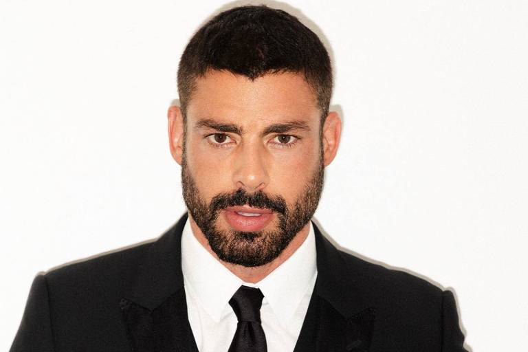 Um homem com cabelo curto e barba bem aparada, vestindo um terno preto com gravata preta e uma camisa branca. Ele está posando de frente para a câmera com uma expressão séria, em um fundo branco.