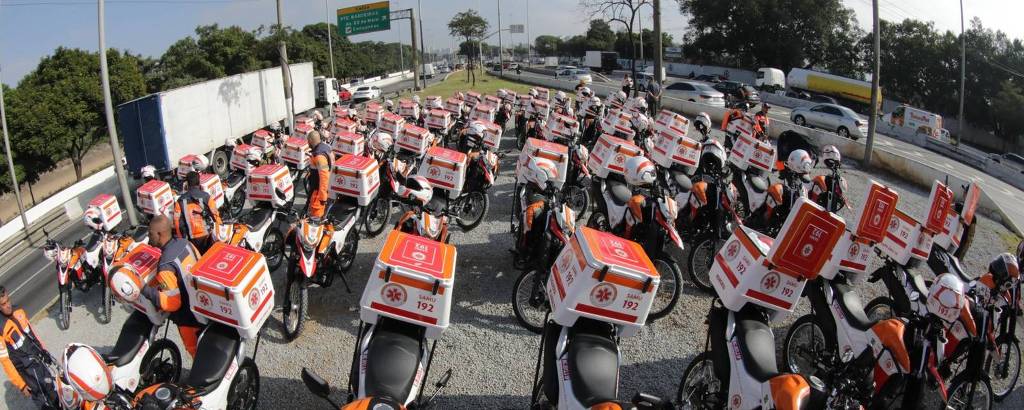 A imagem mostra uma grande quantidade de motocicletas de entrega estacionadas em uma área aberta. As motos são predominantemente brancas e laranjas, com caixas de entrega vermelhas montadas na parte traseira. Ao fundo, é possível ver uma estrada com tráfego e algumas árvores. O céu está claro e há algumas nuvens.
