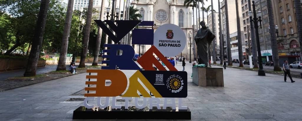 A imagem mostra a Praça da Sé em São Paulo, com a Catedral da Sé ao fundo. Em primeiro plano, há uma estrutura colorida com as palavras 'VERDADE CULTURAL' em destaque. A praça é cercada por palmeiras e edifícios altos, e há algumas pessoas visíveis na área.