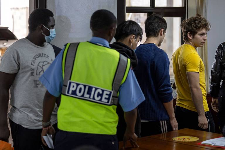 L'immagine mostra un gruppo di persone in un ambiente interno, probabilmente una stanza per interrogatori. Un agente di polizia, che indossa un gilet giallo con la scritta