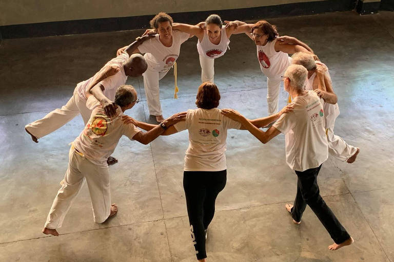 roda de capoeira, com oito pessoas vestidas de branco com braços abertos interligados formando um círculo fechado; A maioria são mulheres