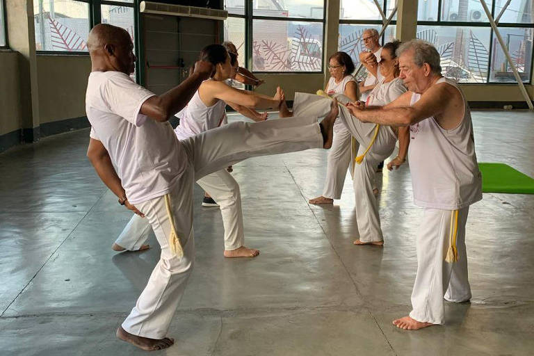 homem negro careca vestido de branco de roupas de capoeira dá chute em outro homem branco em sua frente também com roupas de capoeira, com outros praticantes do esporte ao fundo jogando capoeira