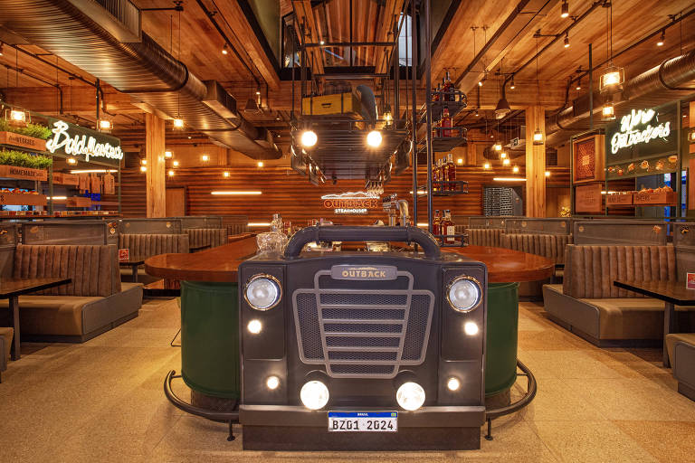 A imagem mostra o interior de um restaurante com um design temático. No centro, há uma estrutura que se assemelha a um caminhão, com a palavra 'OUTBACK' na frente. O ambiente é iluminado com luzes pendentes e possui mesas em formato circular e bancos estofados ao redor. As paredes têm painéis de madeira e há letreiros luminosos com as palavras 'Bold Flavors' e 'Good Times'. O espaço é amplo e bem decorado, criando uma atmosfera acolhedora.
