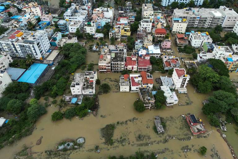A imagem mostra uma vista aérea de uma área urbana afetada por inundações. Várias edificações, predominantemente de vários andares, estão visíveis, com algumas cobertas pela água. O nível da água é alto, e há áreas de vegetação ao redor. Algumas ruas estão submersas, e a água parece turva. O céu está nublado, sugerindo condições climáticas adversas.