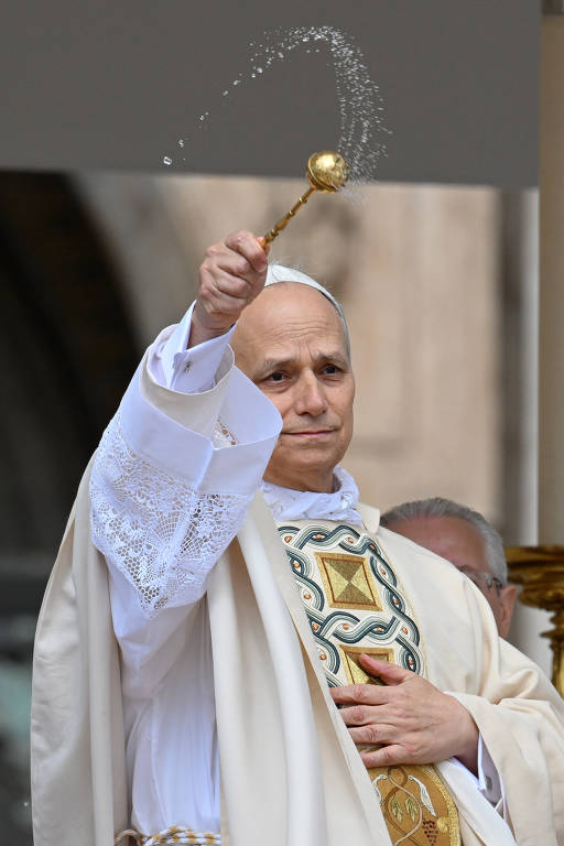 Um homem branco vestido com vestes litúrgicas brancas e um ornamento colorido na parte frontal está fazendo um gesto de bênção.