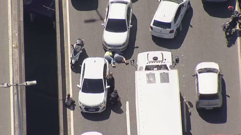 A imagem mostra uma cena de trânsito congestionado, com vários carros parados em uma via. No centro, duas pessoas estão agachadas perto de um carro branco, enquanto outras pessoas observam. Um caminhão branco está estacionado na frente, e há uma moto ao lado. A iluminação sugere que é um dia ensolarado.