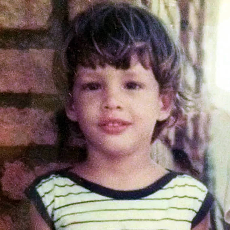 A imagem mostra uma criança com cabelo cacheado e claro, usando uma camiseta listrada em preto e branco. Ela está em frente a uma parede de tijolos e parece estar sorrindo. O fundo é desfocado, com um ambiente que sugere um local interno.