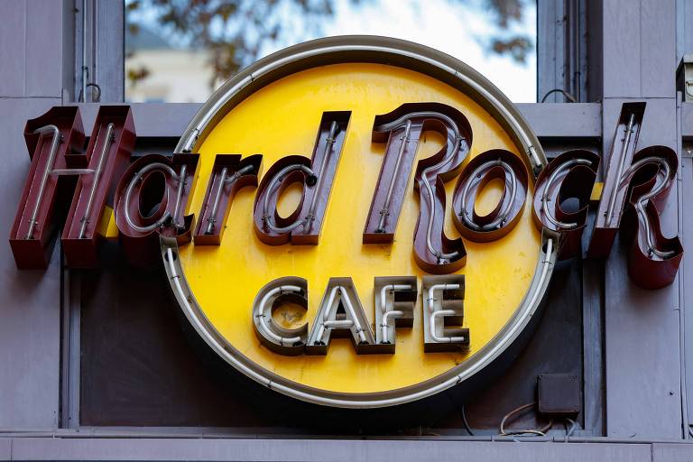 A imagem mostra uma placa do Hard Rock Café, com um fundo amarelo e o nome 'Hard Rock' em letras grandes e escuras, e a palavra 'CAFE' em letras brancas, menores, na parte inferior. A placa é circular e está iluminada, destacando-se em um ambiente urbano.