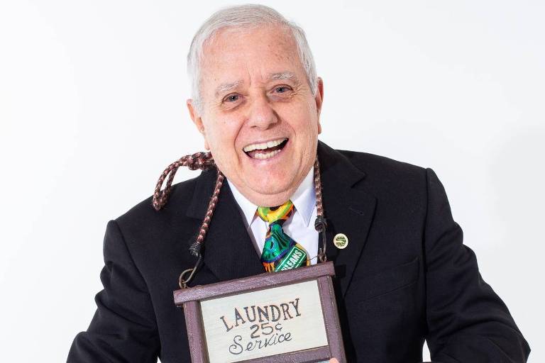 Um homem idoso sorridente está posando para a foto. Ele usa um terno escuro e uma gravata verde. Em suas mãos, ele segura um washboard (tabuleiro de lavar roupas) e um instrumento musical de metal. O fundo da imagem é branco, destacando o homem e os instrumentos que ele segura.