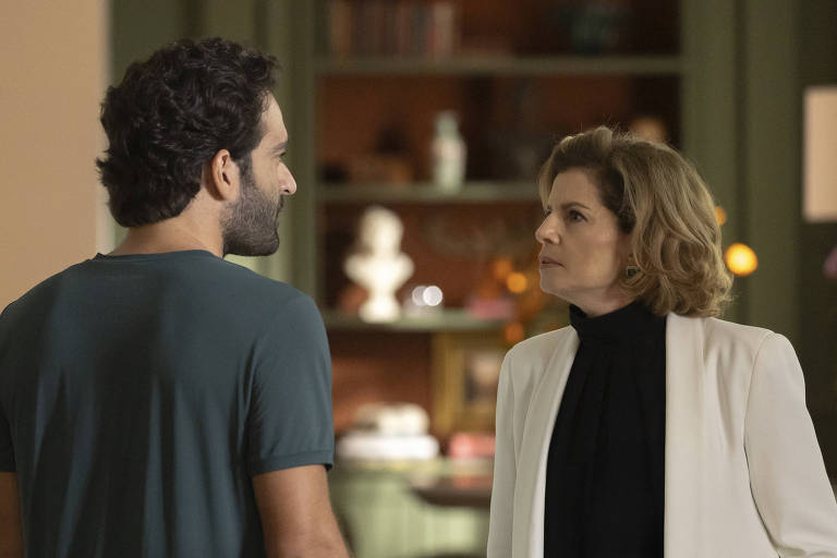 A imagem mostra dois personagens em um ambiente interno. À esquerda, um homem com cabelo cacheado e barba, vestindo uma camiseta azul. À direita, uma mulher com cabelo loiro e ondulado, usando um blazer branco sobre uma blusa preta. Eles estão se encarando, aparentemente em uma conversa séria. Ao fundo, há prateleiras com objetos decorativos e uma iluminação suave.