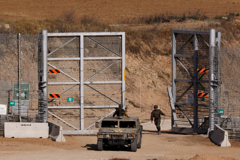 A imagem mostra um portão de segurança militar, com uma estrutura metálica grande e grades. Há um veículo militar tipo HMMWV (Humvee) estacionado à esquerda e um soldado caminhando em direção ao portão. O ambiente ao redor é árido, com vegetação escassa e um terreno seco ao fundo.
