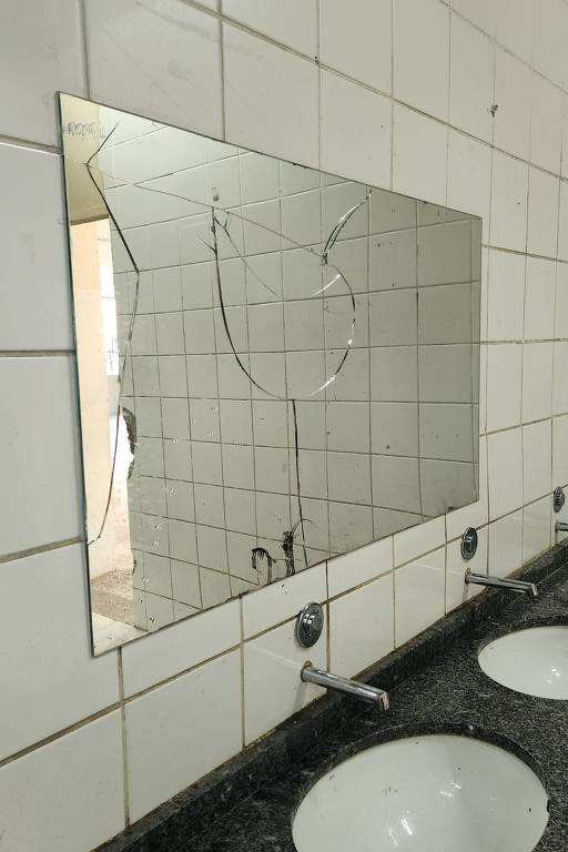 A imagem mostra um espelho grande e quebrado em um banheiro. O espelho está montado em uma parede de azulejos brancos, e a parte quebrada do espelho reflete uma área vazia ao fundo. Abaixo do espelho, há uma bancada de granito escuro com duas pias brancas. O ambiente parece ser um banheiro público.